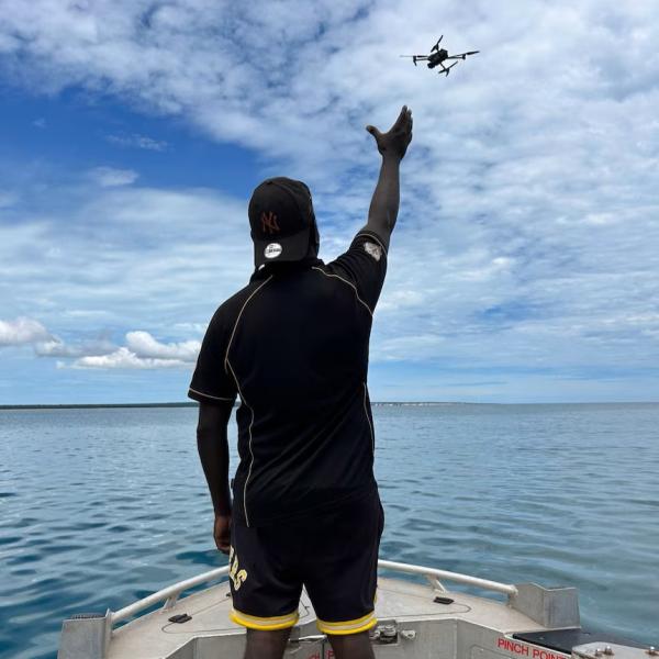 A Tiwi Islander on a boat, arm outstretched towards a drone flying over the ocean.